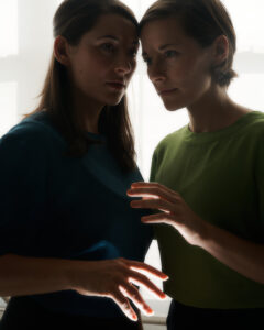two white women in front of a white background, one woman wears a blue top and the other wears a green top; their heads are touching and they're facing each other, each extends an open hand which catches light