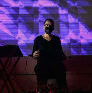 white person wearing black KN95 playing concertina into a computer in front of ageometric purple background in a dark room
