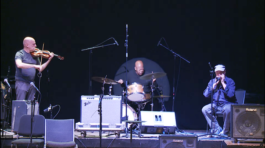 left to right on a stage - a bald white violinist, a bald black drummer, and a bald white harmonica player