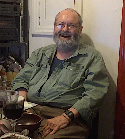 an older white man with glasses and a beard in a green shirt laughing in front of a white wall