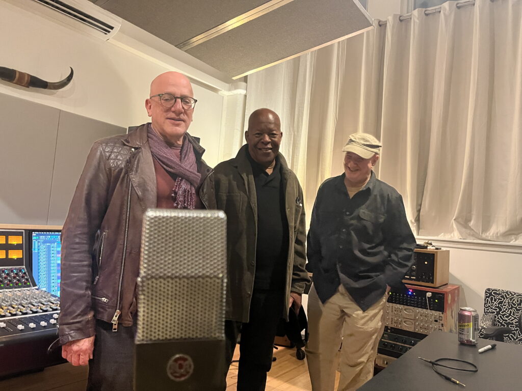 three older men bald men (two white, one black) standing in front of a microphone in a recording studio