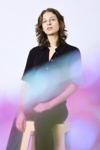 a white woman with shoulder length brown hair wearing black sits on a stool in front of a white wall, pink and blue swirls cover parts of her body